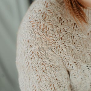 May include: Close-up of a beige knitted sweater with a detailed openwork pattern. The sweater has a crew neck and long sleeves. The intricate design creates a textured look, perfect for fall or winter wear.