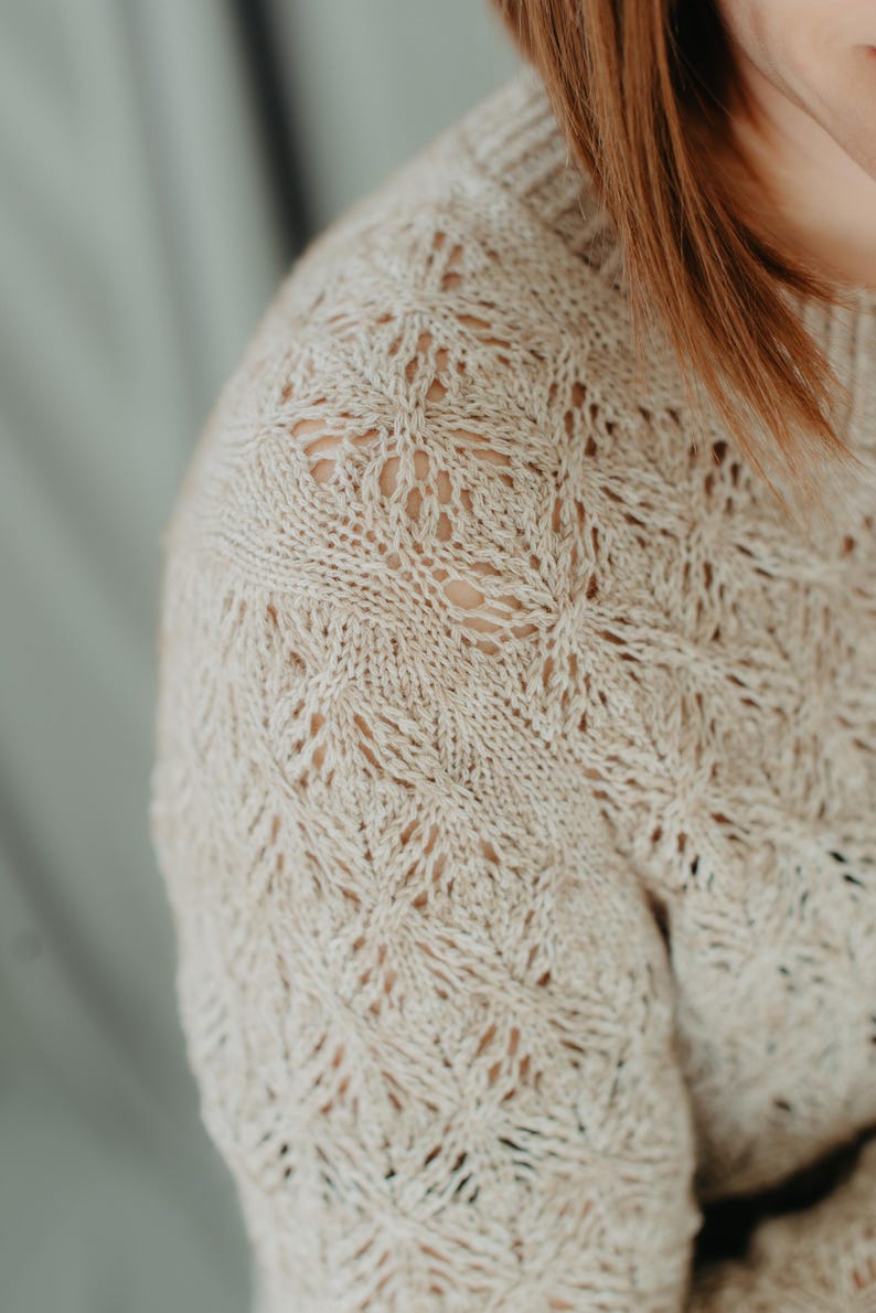 May include: Close-up of a beige knitted sweater with a detailed openwork pattern. The sweater has a crew neck and long sleeves. The intricate design creates a textured look, perfect for fall or winter wear.