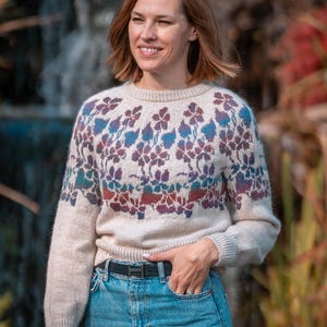 May include: A woman wearing a cream-colored sweater with a floral pattern in shades of purple, blue, and red. She is wearing blue jeans and a black belt.
