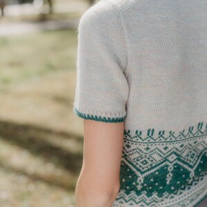 May include: A short-sleeved knit sweater in a light oatmeal color with a speckled texture. The garment features a dark green geometric fair isle pattern around the hem and a matching dark green decorative picot edge on the sleeve cuffs.