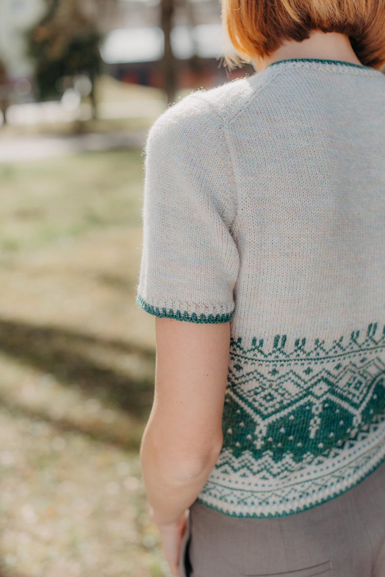 May include: A short-sleeved knit sweater in a light oatmeal color with a speckled texture. The garment features a dark green geometric fair isle pattern around the hem and a matching dark green decorative picot edge on the sleeve cuffs.