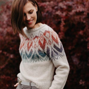 May include: A woman wearing a cream-coloured sweater with a colourful geometric pattern. The sweater has long sleeves and a crew neckline. She is wearing a light grey belt and grey trousers. The background is a blurred image of red foliage.