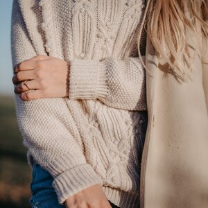 May include: A close-up of a woman wearing a cream-colored cable knit sweater with a textured pattern. The sweater has a loose fit and long sleeves. The woman is also wearing blue jeans and a cream-colored coat.