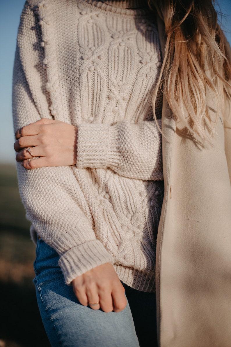 May include: A close-up of a woman wearing a cream-colored cable knit sweater with a textured pattern. The sweater has a loose fit and long sleeves. The woman is also wearing blue jeans and a cream-colored coat.