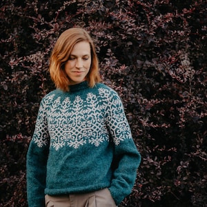 May include: A teal long-sleeve sweater featuring a high neckline and a white intricate geometric snowflake pattern across the chest and shoulders. The sweater is constructed with a ribbed collar, cuffs, and hem, showcasing a textured knit finish.
