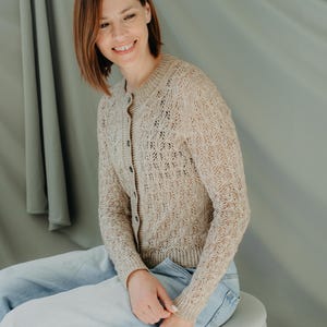 May include: A beige, open-knit cardigan with a button-up front. The cardigan features a delicate, openwork pattern and is paired with light blue jeans. The model is smiling and sitting on a white ottoman.