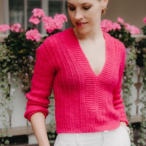 Puede incluir: Una mujer lleva un suéter de punto rosa brillante con un cuello en V profundo y pantalones blancos. Está de pie frente a una pared con flores rosas.