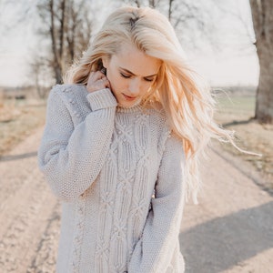 May include: A woman with long blonde hair is wearing a light beige cable knit sweater with a diamond pattern. The sweater has small white pom-poms on the knit.