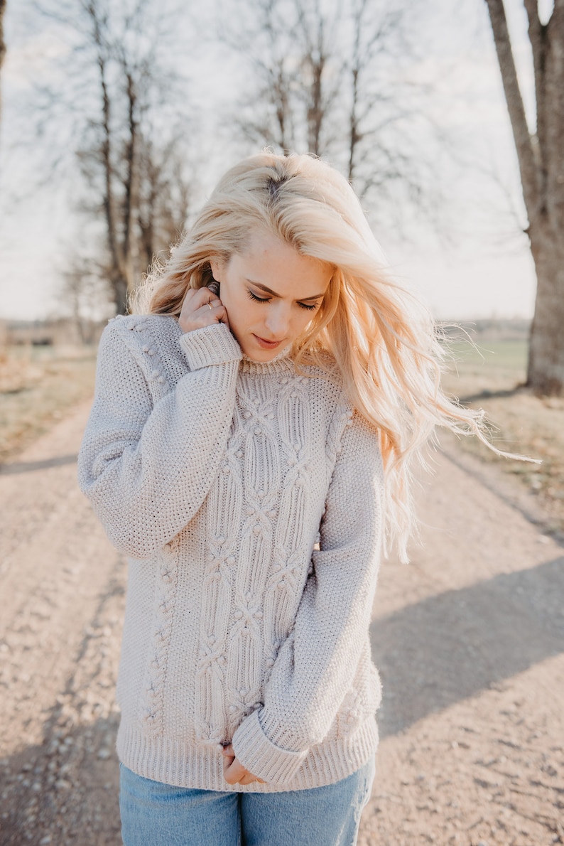 May include: A woman with long blonde hair is wearing a light beige cable knit sweater with a diamond pattern. The sweater has small white pom-poms on the knit.
