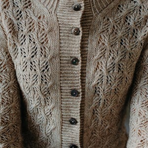 May include: A beige, button-down cardigan sweater with a textured, open-knit design. The sweater features a round neckline and long sleeves. The buttons are a dark brown color. The knit pattern includes diamond and cable designs.