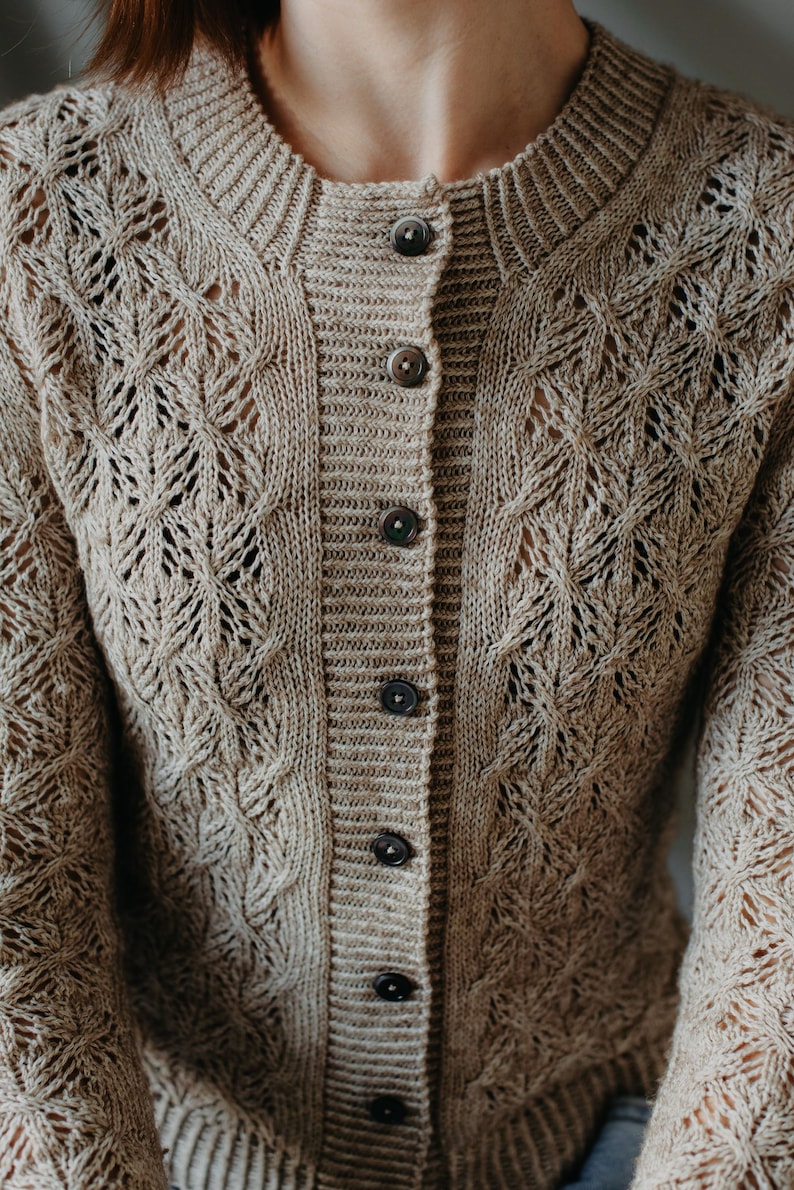 May include: A beige, button-down cardigan sweater with a textured, open-knit design. The sweater features a round neckline and long sleeves. The buttons are a dark brown color. The knit pattern includes diamond and cable designs.