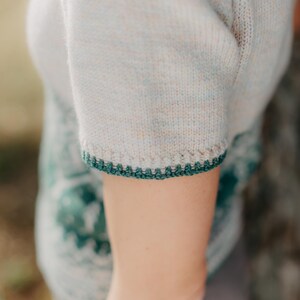 May include: A short-sleeved knit top in a light heathered beige color featuring a dark green crocheted trim along the sleeve edge. The garment displays a textured knit pattern with a subtle speckled appearance and a dark green decorative border.