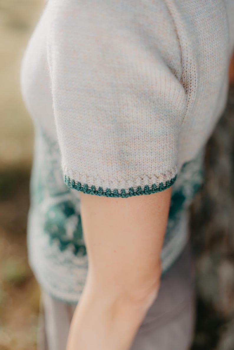 May include: A short-sleeved knit top in a light heathered beige color featuring a dark green crocheted trim along the sleeve edge. The garment displays a textured knit pattern with a subtle speckled appearance and a dark green decorative border.