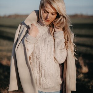 May include: A woman wearing a beige coat over a white cable knit sweater and blue jeans. The woman is standing in a field with green grass and a blue sky in the background.