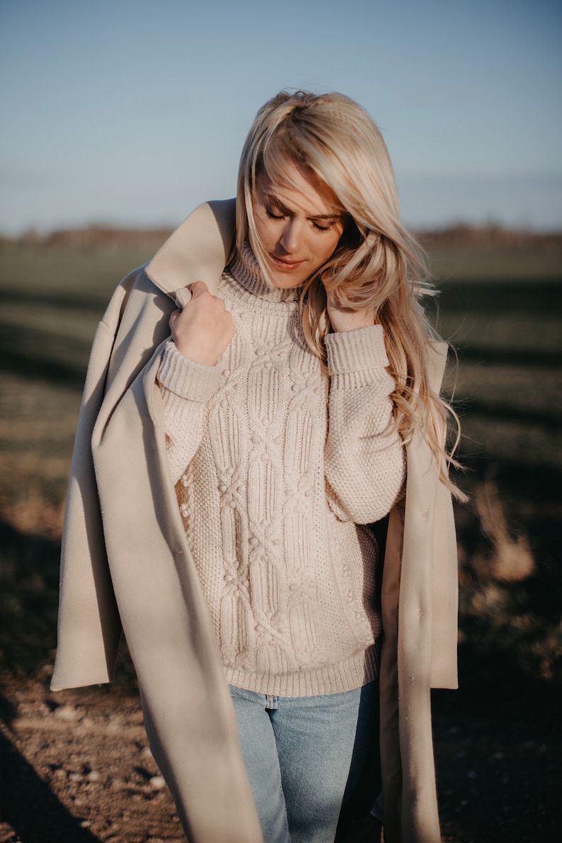 May include: A woman wearing a beige coat over a white cable knit sweater and blue jeans. The woman is standing in a field with green grass and a blue sky in the background.