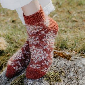 May include: A pair of knitted socks in shades of rust and white, featuring a patterned design. The socks are visible from the ankles down, resting on a moss-covered rock. The socks have a ribbed cuff and a patterned design.
