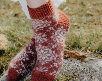 May include: A pair of knitted socks in shades of rust and white, featuring a patterned design. The socks are visible from the ankles down, resting on a moss-covered rock. The socks have a ribbed cuff and a patterned design.