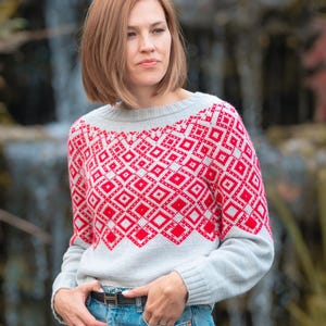 May include: A woman wearing a grey and red patterned sweater with a geometric design. She is wearing blue jeans and a brown belt.