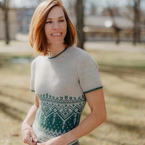 May include: A short-sleeved knit sweater featuring a light beige base color with a dark green geometric pattern across the midsection and waist. The garment includes a round neckline and sleeve cuffs finished with a thin dark green trim detail.
