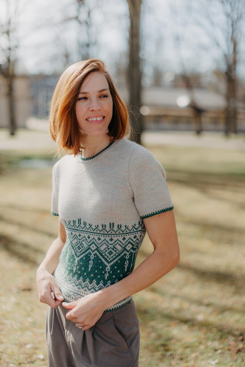 May include: A short-sleeved knit sweater featuring a light beige base color with a dark green geometric pattern across the midsection and waist. The garment includes a round neckline and sleeve cuffs finished with a thin dark green trim detail.