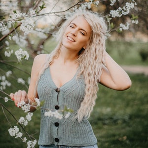 Peut inclure: Une femme aux longs cheveux blonds sourit en se tenant devant un arbre en fleurs. Elle porte un d&eacute;bardeur en tricot bleu clair avec des boutons et un short en jean.