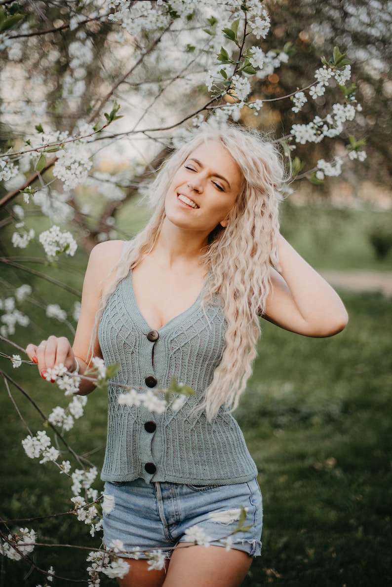 Peut inclure: Une femme aux longs cheveux blonds sourit en se tenant devant un arbre en fleurs. Elle porte un d&eacute;bardeur en tricot bleu clair avec des boutons et un short en jean.