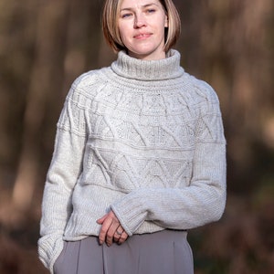 Op de afbeelding: Een vrouw draagt een lichtgrijze trui met een rolkraag en een kabelbreipatroon en een grijze broek. Ze staat in een bosrijke omgeving.