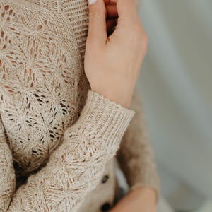 May include: Beige knitted cardigan with a button closure. The cardigan features a detailed openwork pattern on the sleeves and body. The sleeves have ribbed cuffs. The cardigan is made of a soft, textured yarn.