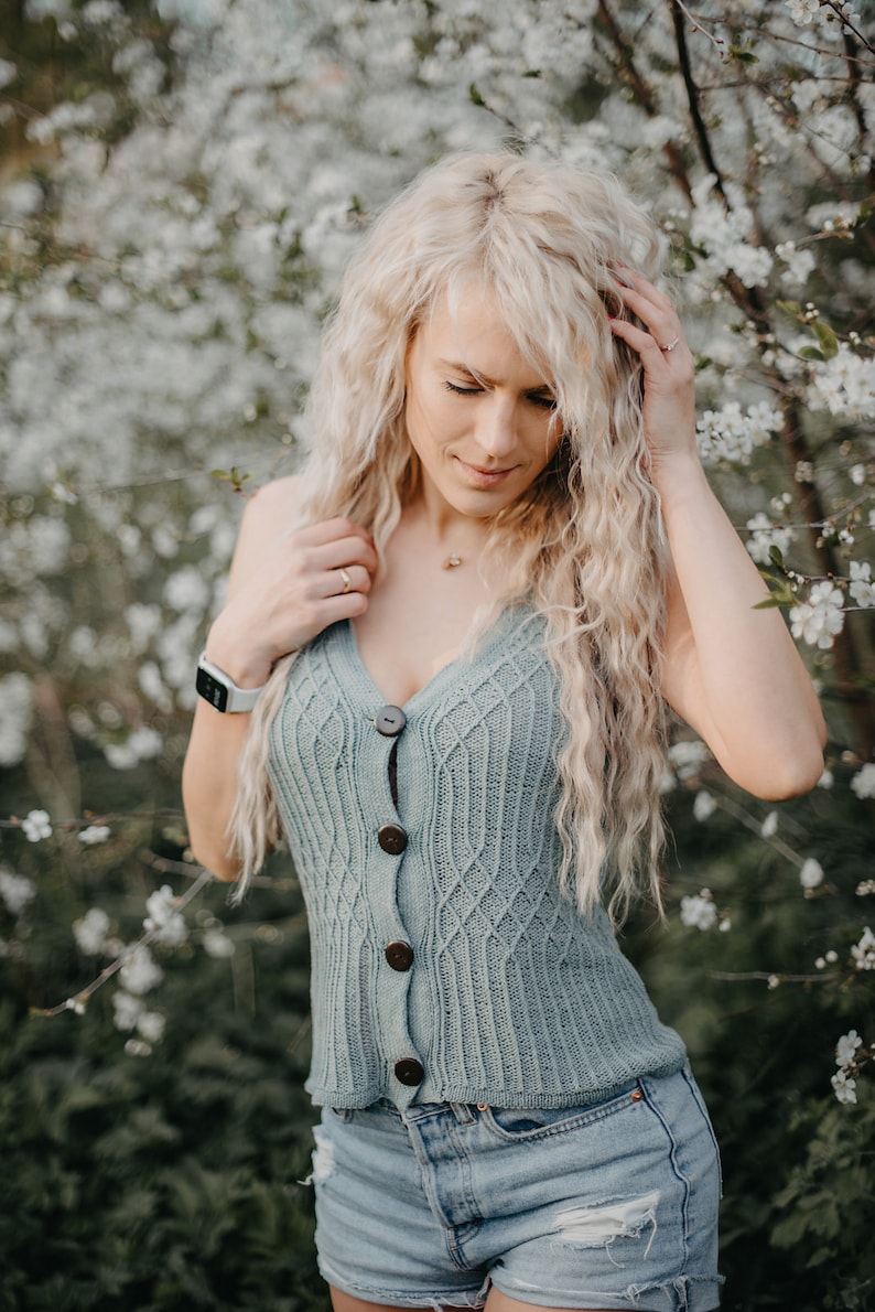 Peut inclure: Une femme aux longs cheveux blonds portant un d&eacute;bardeur en tricot bleu clair avec des boutons en bois et un short en jean. Elle se tient devant un arbre &agrave; fleurs blanches.