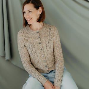 May include: A light brown, button-down cardigan with a delicate open-knit pattern. The long-sleeved sweater has a round neckline and is paired with light blue jeans. The cardigan features small, round buttons and a textured design.