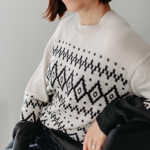 May include: A woman wearing a white jumper with a black and white geometric pattern. The jumper has a crew neck and long sleeves. The woman is sitting in a chair with her hands resting on her lap.