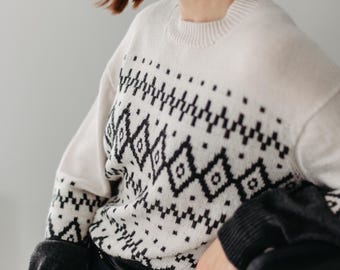 Op de afbeelding: Een vrouw draagt een witte trui met een zwart-wit geometrisch patroon. De trui heeft een ronde hals en lange mouwen. De vrouw zit op een stoel met haar handen op haar schoot.