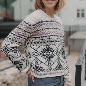 Pode incluir: Camisola creme com um padrão geométrico em preto, roxo e cinza. A camisola tem gola redonda e mangas compridas. O design inclui formas de diamante e ziguezague. A camisola é usada com jeans azuis.