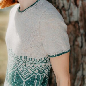 May include: A short-sleeved knit sweater featuring a cream-colored upper section and a dark green lower section with a detailed geometric pattern. The garment has a round neckline and sleeve cuffs finished with a dark green decorative trim.