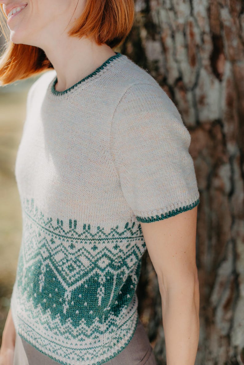 May include: A short-sleeved knit sweater featuring a cream-colored upper section and a dark green lower section with a detailed geometric pattern. The garment has a round neckline and sleeve cuffs finished with a dark green decorative trim.
