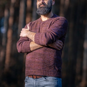 May include: A man with a shaved head and a full beard is wearing a maroon sweater and blue jeans. He is standing in front of a blurred background of trees.