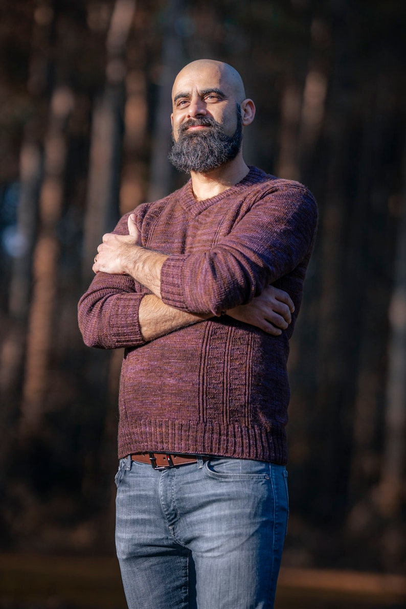 May include: A man with a shaved head and a full beard is wearing a maroon sweater and blue jeans. He is standing in front of a blurred background of trees.