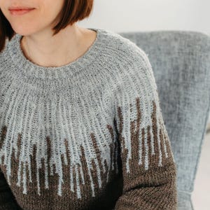 Op de afbeelding: Een close-up van een grijze en bruine gebreide trui met een verticaal gestreept patroon. De trui heeft een ronde hals en lange mouwen.