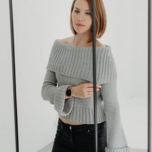 Può includere: Un maglione grigio con spalle scoperte e maniche a campana. Il maglione ha una trama a coste ed è abbinato a jeans neri. La modella indossa un orologio al polso.