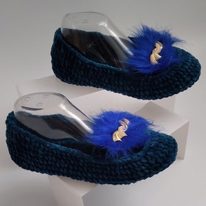 Może przedstawiać: Para zielonych aksamitnych kapci z niebieskimi, puszystymi pomponami i złotymi akcentami.