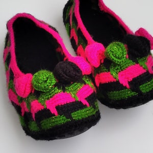 Może przedstawiać: Para czarnych, zielonych i różowych wełnianych kapci z pomponami. Kapcie mają kratkowany wzór.
