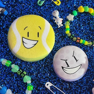 Puede incluir: Una colección de accesorios divertidos sobre una superficie azul. Incluye un botón amarillo con forma de pelota de tenis con una cara sonriente, un botón con forma de pelota de golf lila claro, pulseras de cuentas, imperdibles y una pequeña figura.