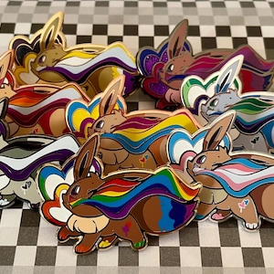 May include: A set of ten enamel pins featuring a cartoon character with different colored pride flags. The character is brown with a long, pointed snout and large ears. The flags are rainbow, transgender, bisexual, lesbian, pansexual, asexual, genderfluid, non-binary, intersex, and aromantic.