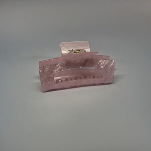 Könnte beinhalten: Ein rosa Plastik-Haarklammer mit strukturierter Oberfläche. Die Klammer hat eine gebogene Form und einen federbelasteten Mechanismus.