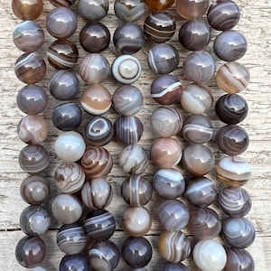 Könnte beinhalten: Eine Kette aus grauen und weißen Achatperlen mit braunen und weißen Bändern. Die Perlen sind rund und glatt und auf einer durchsichtigen Gummikordel aufgefädelt.