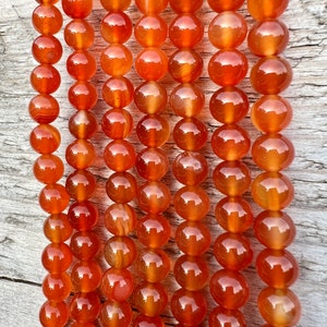 Könnte beinhalten: Eine Strähne aus Karneolperlen in verschiedenen Orangetönen und Rottönen. Die Perlen sind rund und glatt und haben eine natürliche, polierte Oberfläche.