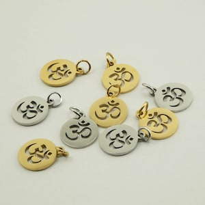 May include: A collection of gold and silver-colored Om symbol pendants. The pendants are circular with a cutout design of the Om symbol. Each pendant has a small loop for attaching to a chain or bracelet. The pendants are displayed on a light-colored surface.