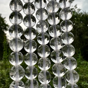 May include: A strand of clear quartz crystal beads, each bead is round and smooth. The beads are strung on a white thread and are hanging against a blurred background of green foliage.