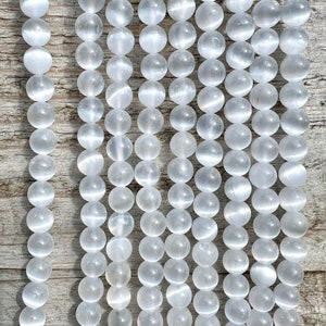 Selenite gemstone strands laid on a pieces of wood in outdoor and photographed under natural light. These beads are semi transparent with some white hues. Selenite beads have chatoyancy effect.