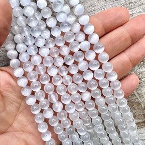 Selenite gemstone 8mm beads strands held in hand on a pieces of wood in outdoor and photographed under natural light. These beads are semi transparent with some white hues. Selenite beads have chatoyancy effect.